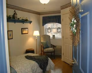 A small bedroom with a single bed covered in a light-colored bedspread and a dark throw blanket. There is a wooden chair with a cushion near a window with blue curtains. A floor lamp is next to the chair, and a small shelf with decorative items is mounted on the wall. The door is open and has a wreath hanging on it.
