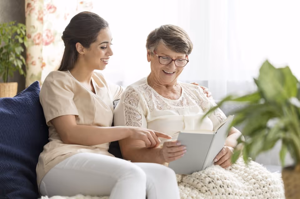 A young caregiver sitting on a couch next to an elderly woman, both smiling and looking at a book together in a bright, cozy living room with plants and floral curtains.