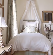 Single bed with white bedding and a sheer canopy, flanked by bedside tables, lamps, a mirror, and floral curtains in a cozy bedroom.
