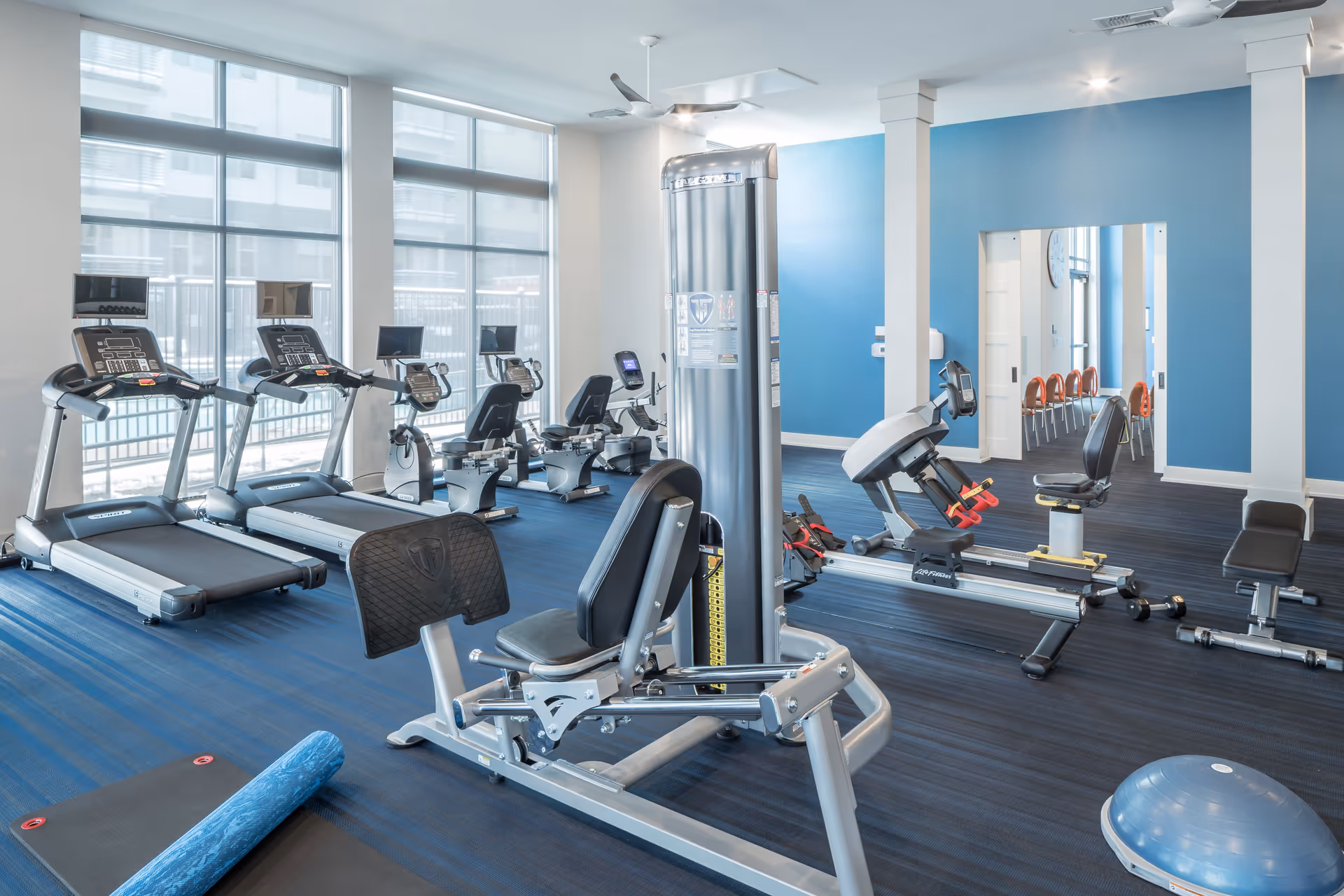 A bright fitness room with various exercise equipment including treadmills, stationary bikes, weight machines, a yoga mat with a foam roller, and a balance trainer. The room has large windows allowing natural light to enter, blue walls, and dark blue flooring.