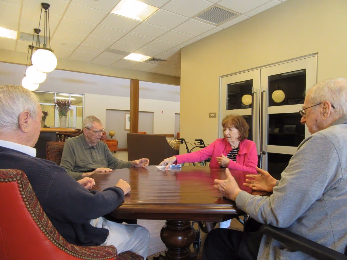 Four elderly people sitting around a wooden table in a well-lit room with beige walls and ceiling lights. One woman in a pink jacket is reaching across the table, possibly handing something to one of the men. The room has comfortable chairs and a cozy atmosphere.