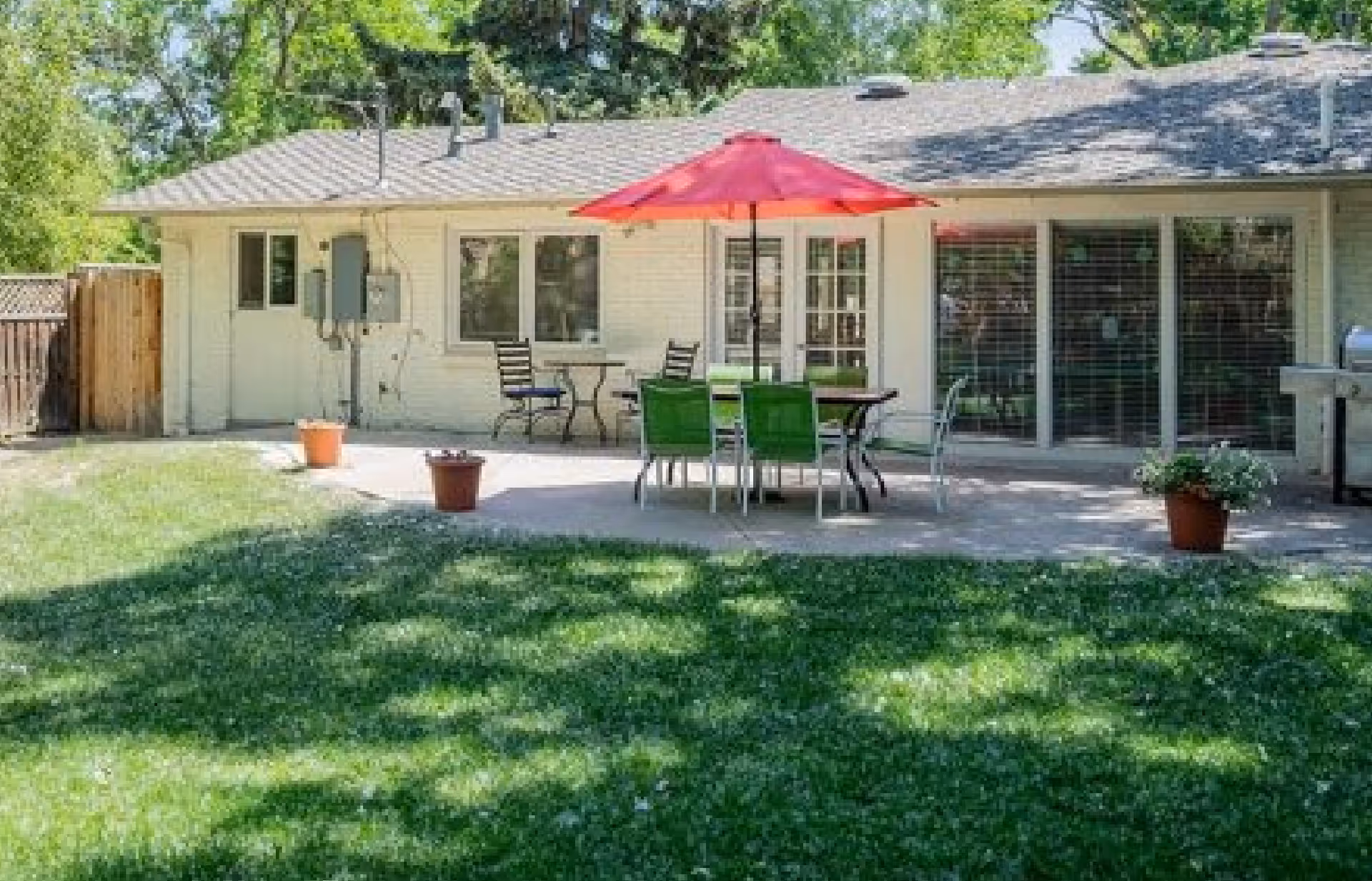 Outdoor patio area of a single-story building with a red umbrella over a table surrounded by green and black chairs. There are potted plants on the patio and a grassy lawn in the foreground. The building has multiple windows and a sliding glass door.