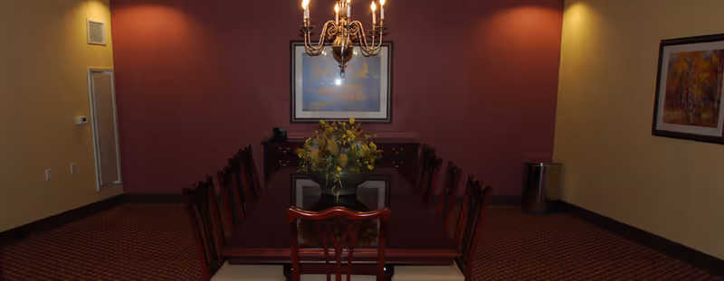 A formal dining room with a long wooden table surrounded by chairs. A chandelier hangs above the table, and a floral centerpiece is placed in the middle. The walls are painted in warm tones with framed artwork, and there is a sideboard against the far wall.