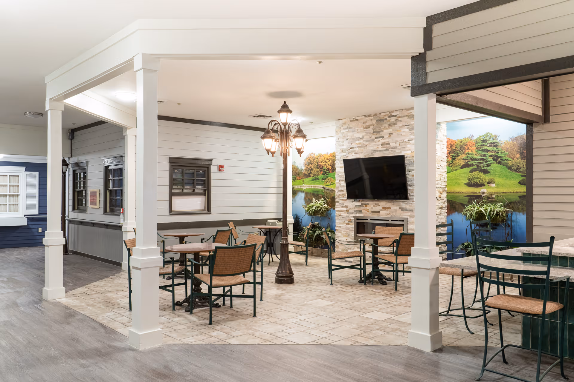 Indoor common area with several tables and chairs arranged around a central lamp post. The space features a stone fireplace with a mounted flat-screen TV above it. The walls have large scenic murals depicting a green landscape with trees and a pond. The flooring is a mix of tile and wood-like material.