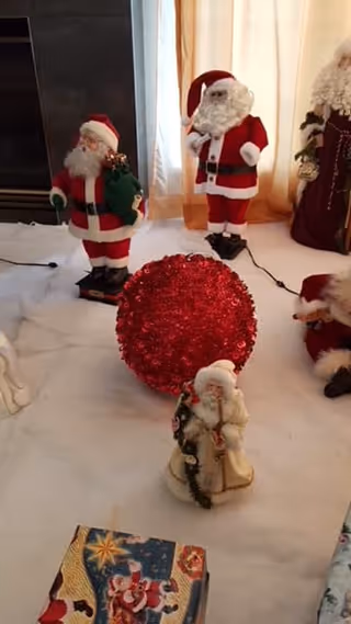A festive indoor scene with several Santa Claus figurines and a large red glittery ornament placed on a white surface, with a curtain and part of a fireplace visible in the background.