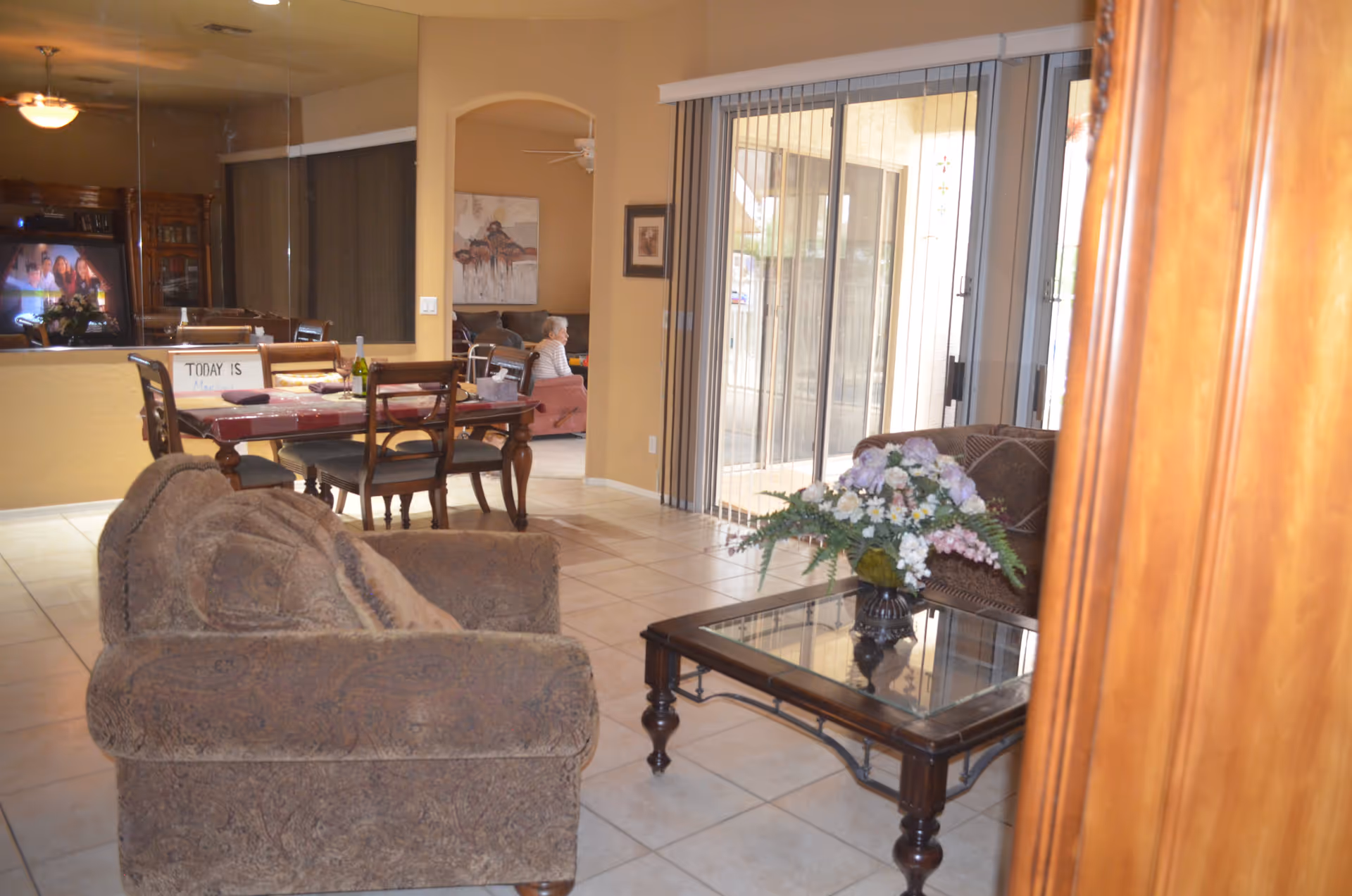 Interior view of a living room and dining area in an assisted living home. The living room has a patterned armchair and a glass coffee table with a floral arrangement. The dining area has a wooden table with chairs and a sign that says 'TODAY IS Monday'. A person is sitting on a couch in the background near a large sliding glass door with vertical blinds.