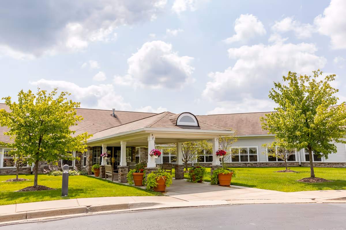 Front exterior view of Independence Village of Oxford (Waterstone) showing a covered entrance with white pillars, stone bases, hanging flower baskets, potted plants, benches, green lawn, and trees under a partly cloudy sky.