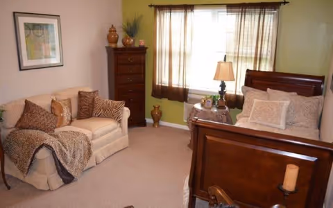 A cozy bedroom featuring a wooden bed with beige and patterned pillows, a small wooden side table with a lamp and decorative items, a beige loveseat with patterned cushions and a throw blanket, a tall wooden dresser with decorative vases on top, and a window with brown curtains allowing natural light into the room.