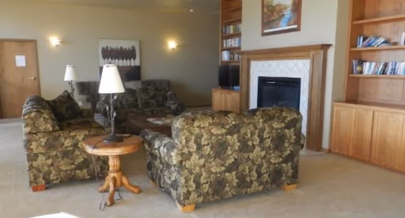A carpeted common living room with floral-patterned sofas arranged around a fireplace, wooden bookshelves, and side tables with lamps.