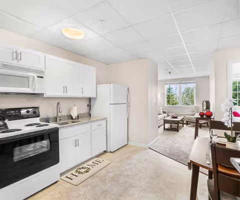 A bright kitchen area with white cabinets, a white refrigerator, a microwave, and a black stove. The kitchen has a granite countertop with a sink and a paper towel holder. A beige mat with the word 'HOME' is placed on the floor. Beyond the kitchen, there is a living room with a large window, a sofa, a coffee table, and a side table with decorative items.