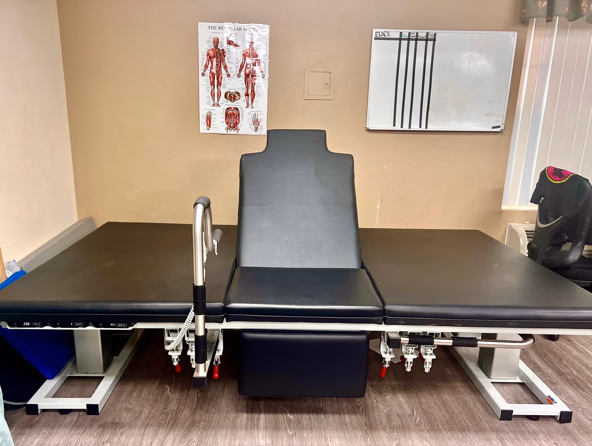 A medical examination or therapy table with black padding in a room with beige walls. On the wall behind the table, there is a poster of the muscular system and a whiteboard with some writing. A black office chair is partially visible to the right near a window with vertical blinds.