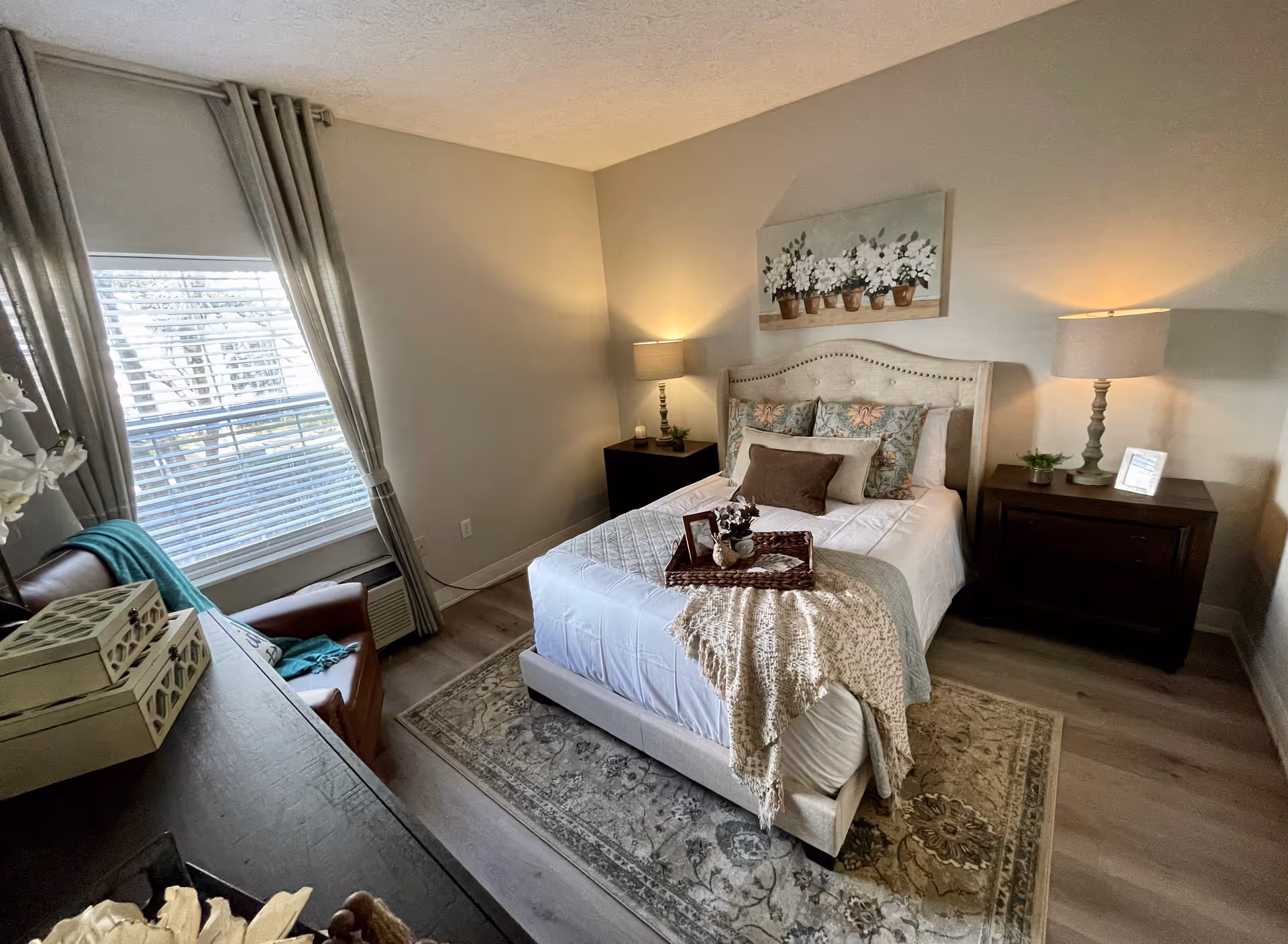 A cozy bedroom with a neatly made bed featuring multiple pillows and a throw blanket. There are two bedside tables with lamps on each side of the bed. A window with curtains lets in natural light, and a comfortable chair with a teal throw is positioned near the window. The room has a soft, neutral color palette with a floral painting above the bed and a patterned rug on the floor.