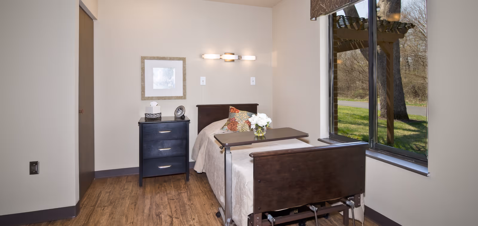 A small, neatly arranged bedroom in a senior living facility with a single bed featuring a dark wooden headboard and footboard. The bed is covered with a beige blanket and has a colorful pillow and a vase of white flowers on an overbed table. Next to the bed is a black three-drawer nightstand with a tissue box and a decorative clock. Above the nightstand is a framed picture and a wall-mounted light fixture. A large window on the right side shows an outdoor view with trees and grass.