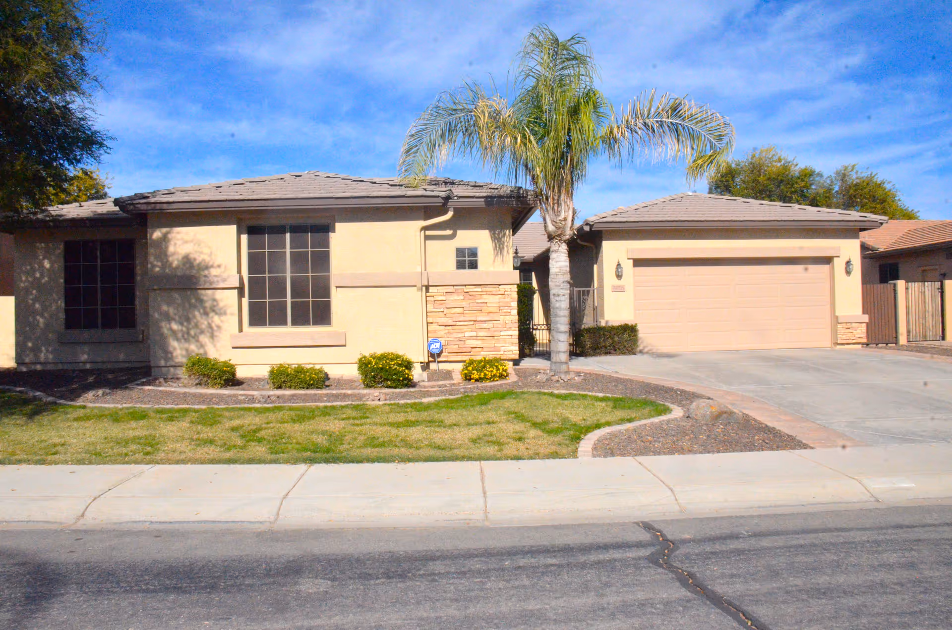 Single-story residential building with beige exterior walls, a tiled roof, a large window, a garage door, a palm tree in the front yard, and a well-maintained lawn and shrubs under a blue sky.