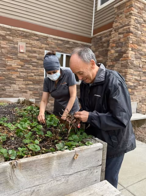 Two people tending to plants in a raised garden bed outside a building with stone and siding exterior walls. One person is wearing a face mask and head covering, while the other is smiling and pulling weeds or plants.