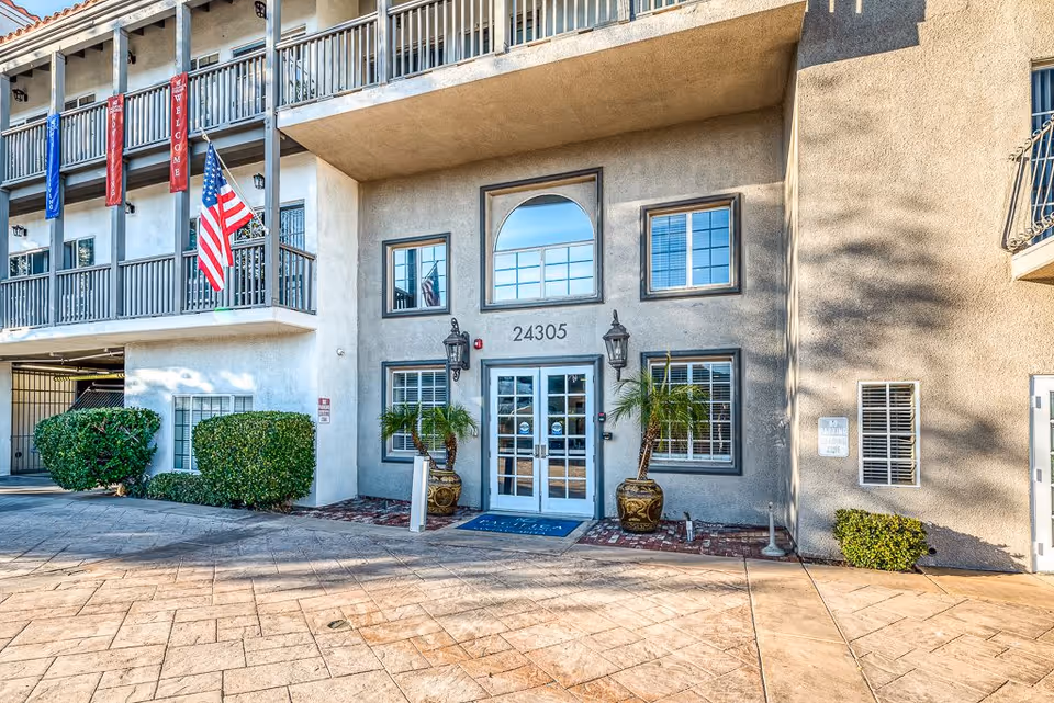 Exterior entrance of a senior living facility with the address number 24305 above double glass doors. Two potted plants flank the entrance, and an American flag is displayed on the left side. The building has balconies with railings and banners that say 'Welcome'.