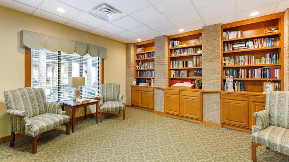 A cozy reading room with three patterned armchairs arranged around a small wooden table with a lamp and a book. The room features large built-in wooden bookshelves filled with books and decorative items. A large window with blinds and a valance lets in natural light.