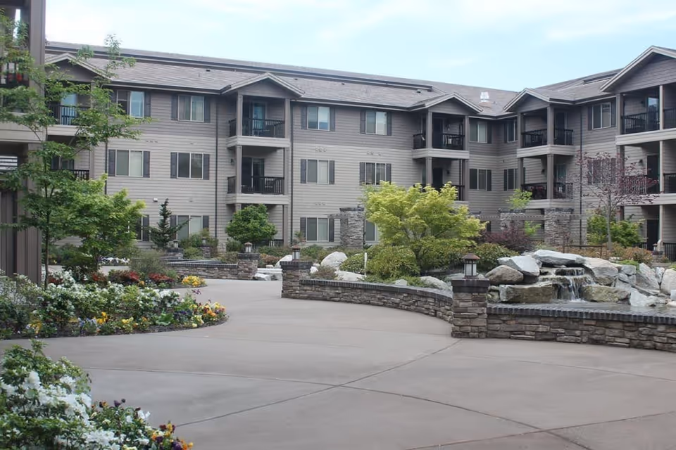 Outdoor courtyard area of The Springs at Veranda Park senior living facility featuring a paved walkway, landscaped garden beds with flowers and shrubs, a stone water feature with a small waterfall, and a multi-story residential building with balconies in the background.