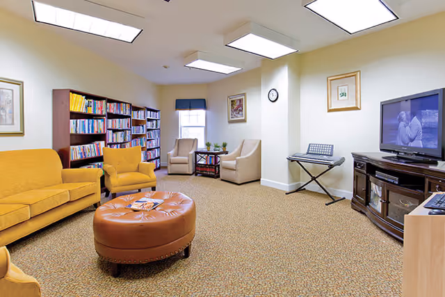 A cozy living room area with mustard yellow and beige armchairs and a sofa, a round brown leather ottoman in the center, bookshelves filled with books along one wall, a flat-screen TV on a dark wooden stand, a keyboard on a stand, and framed artwork on the walls. The room is well-lit with ceiling lights and has a window with a dark valance.