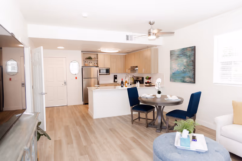 Bright open-plan living area with a small round dining table and blue chairs, kitchen island, and stainless steel appliances.