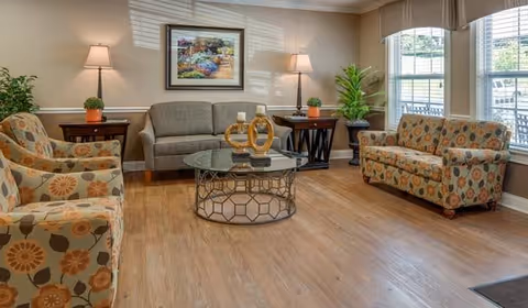 A cozy living room with two patterned armchairs, a gray sofa, a glass coffee table with decorative items, two side tables with lamps, a framed floral painting on the wall, and large windows letting in natural light.