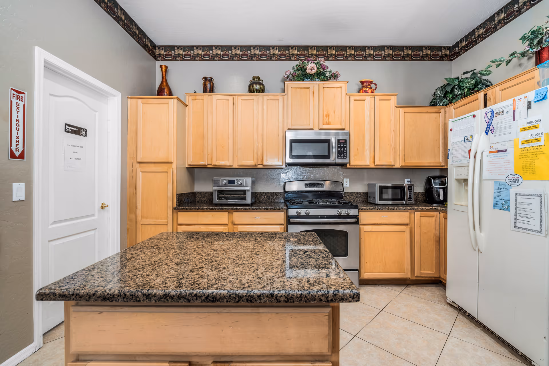 A kitchen with light wood cabinets, a granite countertop island in the center, stainless steel oven and microwave, a toaster oven, a microwave, a coffee maker, and a white refrigerator covered with various papers and notes. There is a door labeled 'Employees Only' and a fire extinguisher sign on the left wall. Decorative vases and plants are placed on top of the cabinets.