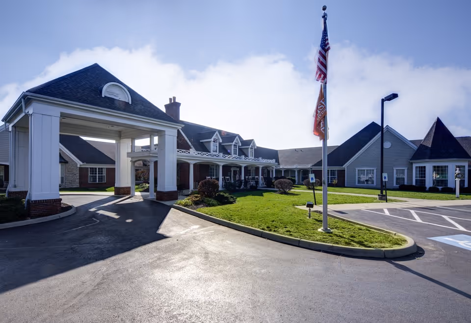Exterior view of Sunrise of Findlay senior living facility showing a covered entrance driveway, manicured lawn, American flag, and multiple connected buildings with various roof styles under a partly cloudy sky.