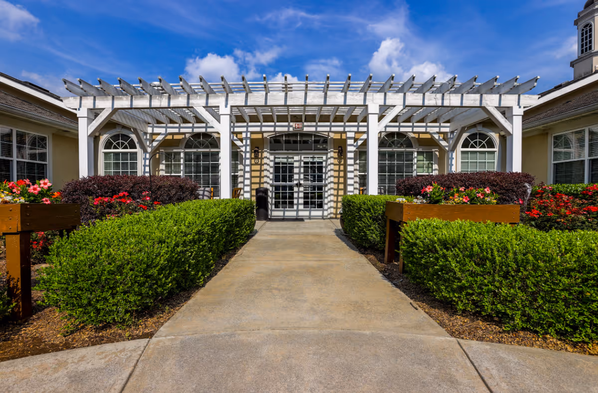 Entrance to a senior living facility with a white pergola structure over the walkway, surrounded by neatly trimmed green bushes and colorful flowers under a bright blue sky with some clouds.