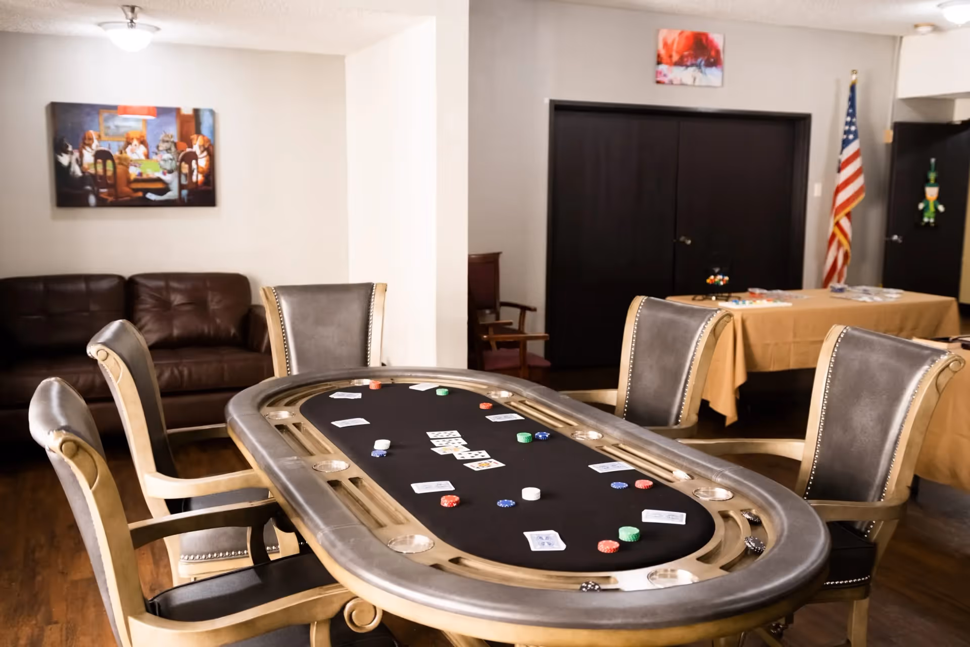 A game room with a poker table featuring playing cards and poker chips. The room has four leather chairs around the table, a brown leather couch against the wall, an American flag in the corner, and a table covered with a beige cloth in the background. There are two paintings on the walls and wooden flooring.