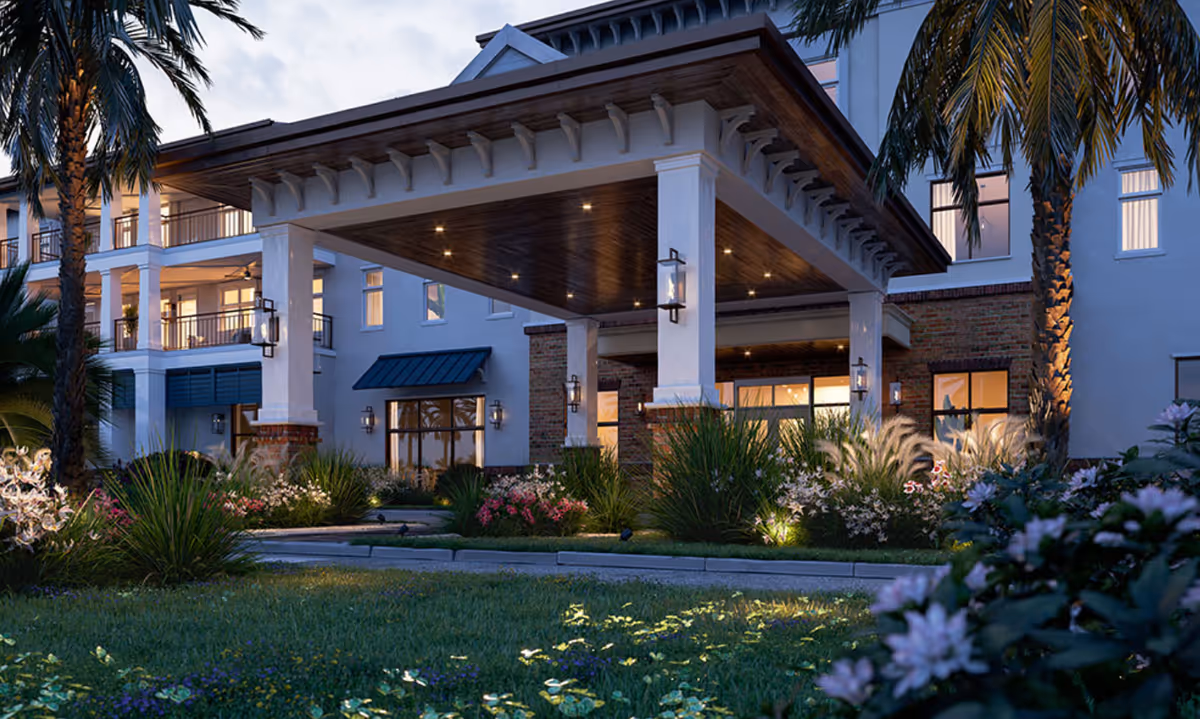 Exterior view of Seafields at Kiawah Island facility entrance at dusk, featuring a covered drop-off area with columns, warm lighting, palm trees, and landscaped garden beds with various plants and flowers.