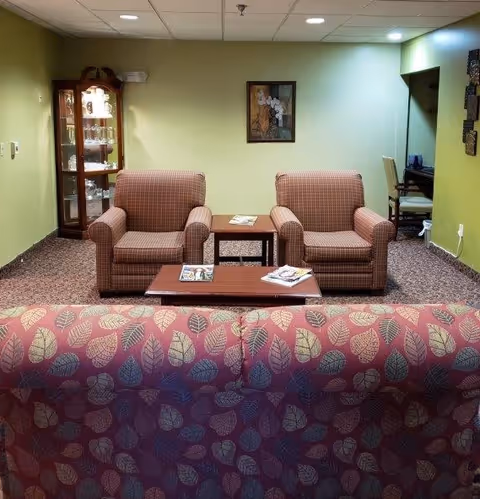 A cozy living room area with two patterned armchairs and a matching sofa with a leaf design. There are two wooden tables with magazines on them, a glass display cabinet in the corner, a framed picture on the wall, and a small desk with a chair in a recessed area.