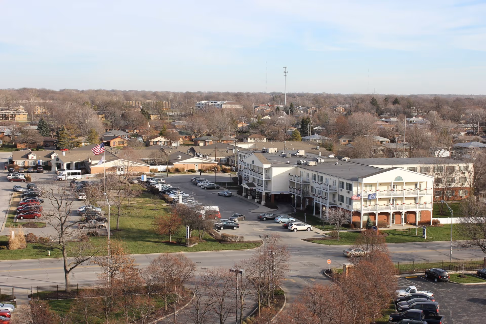 Aerial view of a three-story senior living facility with balconies, parking areas, a flagpole, and surrounding neighborhood.