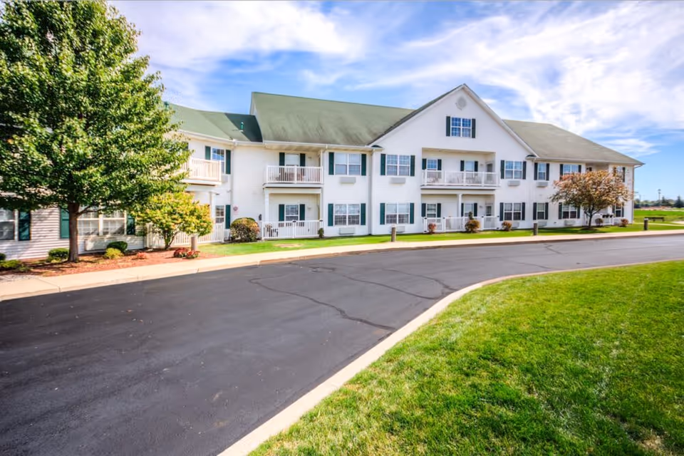 Front exterior of a two-story white senior living building with balconies, a curved driveway, lawn, and trees.