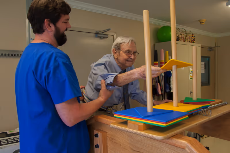 An elderly man wearing glasses and a light blue shirt is engaged in a physical therapy activity involving stacking colorful square boards on wooden poles, assisted by a caregiver in blue scrubs inside a room with therapy equipment.