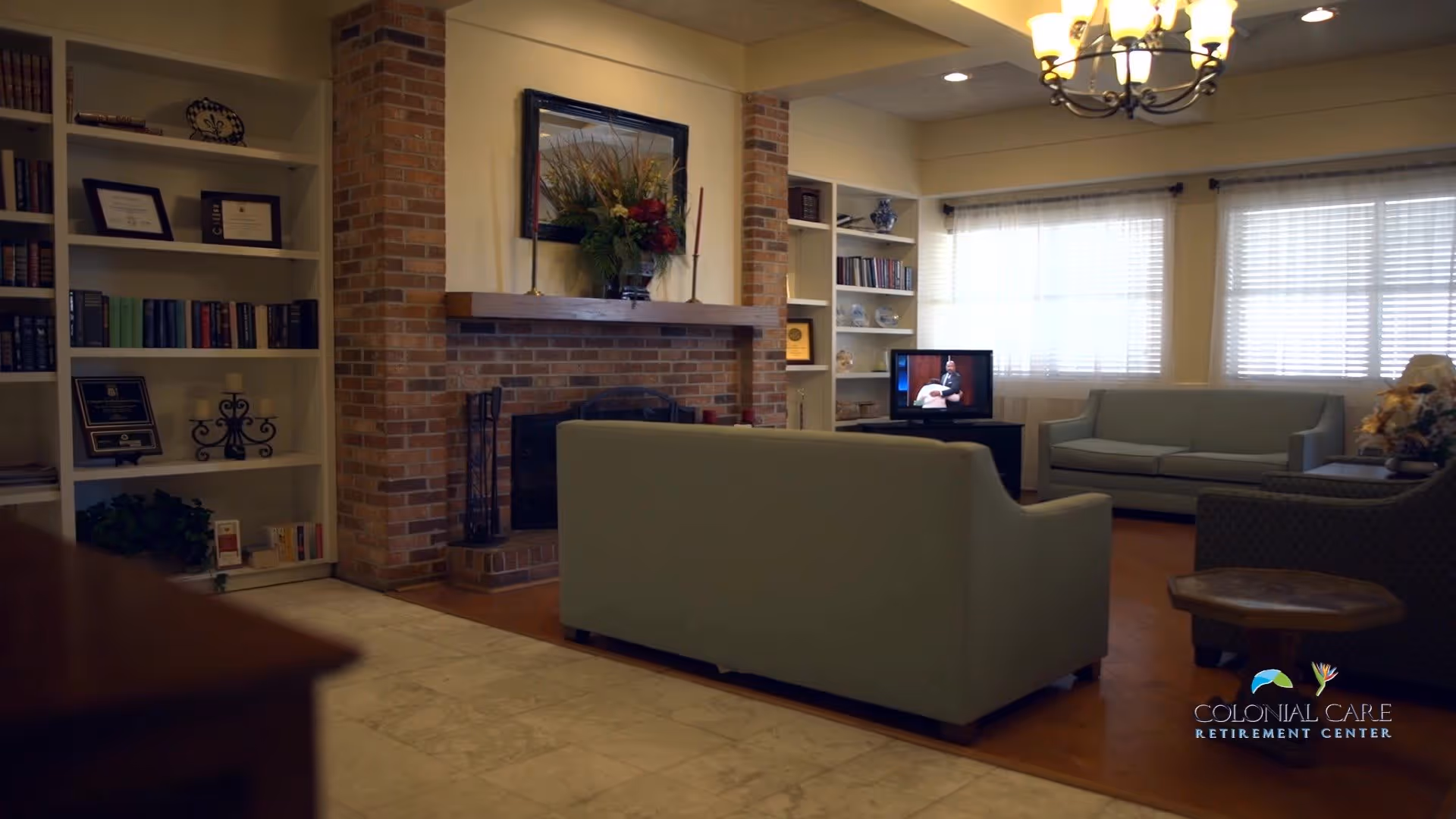 A cozy living room with two light green sofas, a patterned armchair, a wooden coffee table, a brick fireplace with a wooden mantle decorated with a floral arrangement, built-in white bookshelves filled with books and decorative items, a television on a stand, and large windows with white blinds letting in natural light. A chandelier hangs from the ceiling.