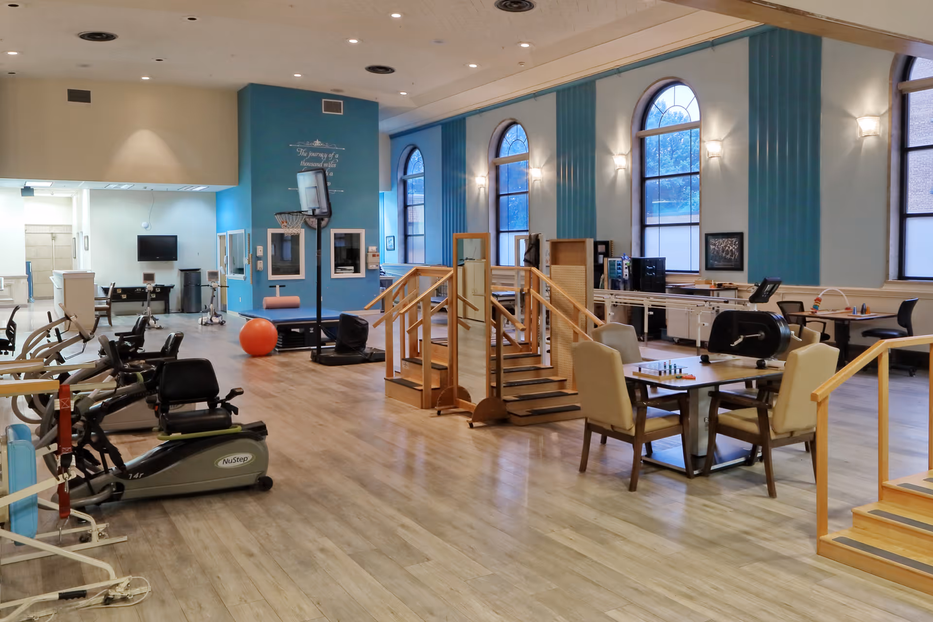 A spacious rehabilitation or therapy room with exercise equipment, parallel bars, wooden stairs for physical therapy, tables and chairs, and large arched windows letting in natural light.