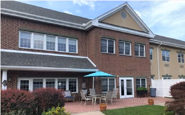 Exterior view of a two-story brick and beige building with multiple windows and a patio area. The patio has a table with six chairs and a blue umbrella. There are potted plants and bushes surrounding the patio, and a white fence is visible on the right side. The sky is partly cloudy.