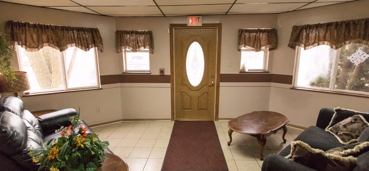 A cozy living room area with two dark leather sofas facing each other, a wooden coffee table in the center, and a small table with a flower arrangement. The room has tiled floors, three windows with brown valance curtains, and a wooden front door with an oval glass panel. An exit sign is visible above the door.
