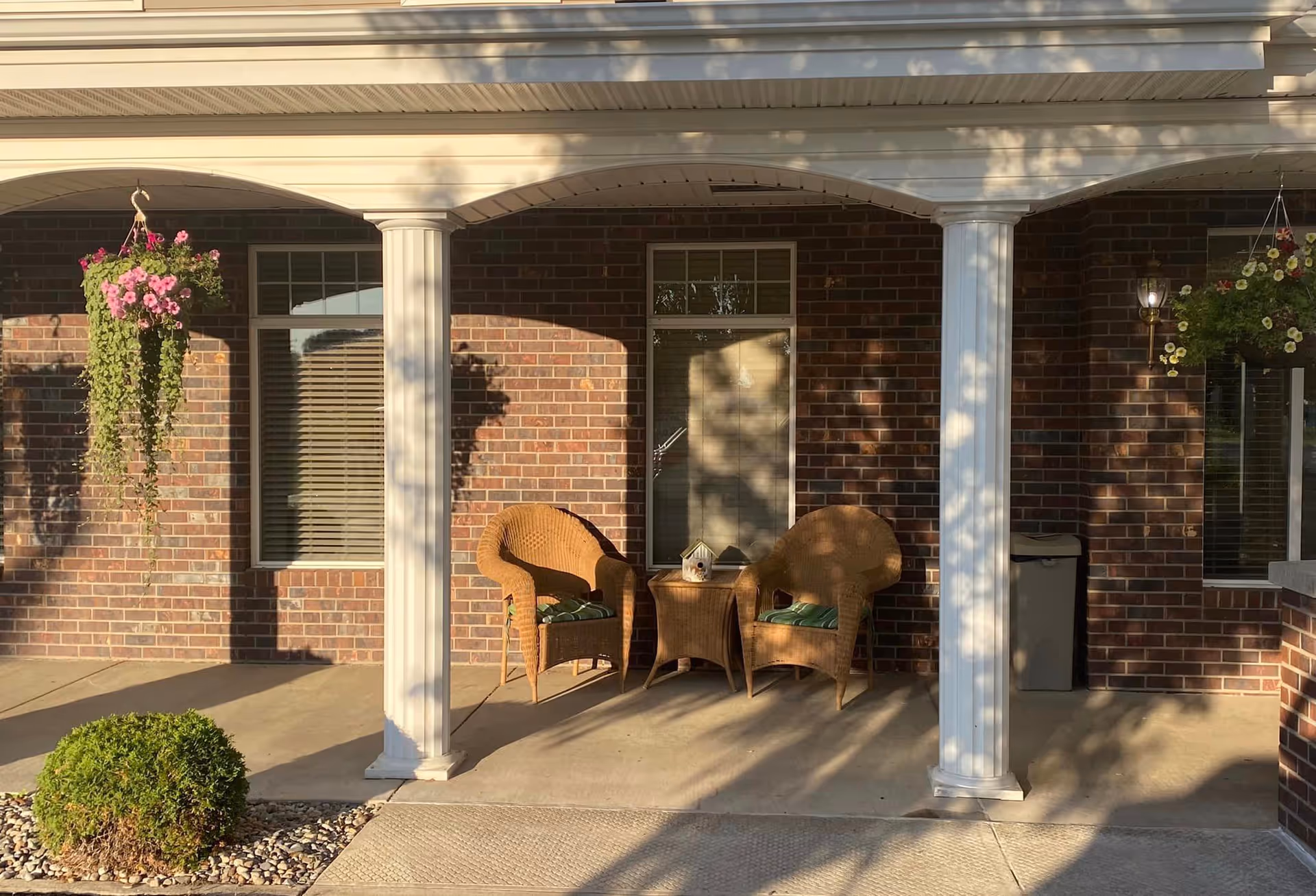 A covered outdoor patio area with two wicker chairs and a small wicker table between them, placed against a brick wall with two windows. There are white columns supporting the roof, hanging flower baskets on either side, and a small green bush in the foreground.