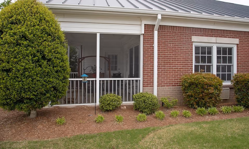 Exterior view of a brick building with a screened-in porch area. The porch has white railings and a swing inside. There are neatly trimmed bushes and a small tree in front of the building, with a well-maintained lawn and mulch bed.