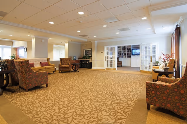 Spacious and well-lit common area with patterned carpet, multiple upholstered chairs, a sofa, side tables with decorative items, and French doors leading to a room with bookshelves and a television.