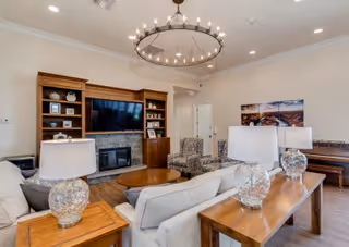 A cozy living room with a large flat-screen TV mounted on a wooden entertainment center above a stone fireplace. The room features a beige sofa, two patterned armchairs, a round wooden coffee table, and a wooden console table with three glass lamps. The walls are light-colored, and there is a circular chandelier hanging from the ceiling. A piano and a landscape painting are visible in the background.
