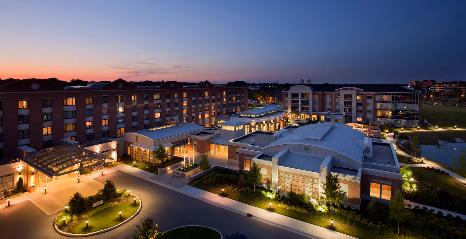 Aerial view of the Marquette senior living facility at dusk, showing a large multi-story building with illuminated windows and exterior lights, surrounded by landscaped grounds and a driveway entrance.
