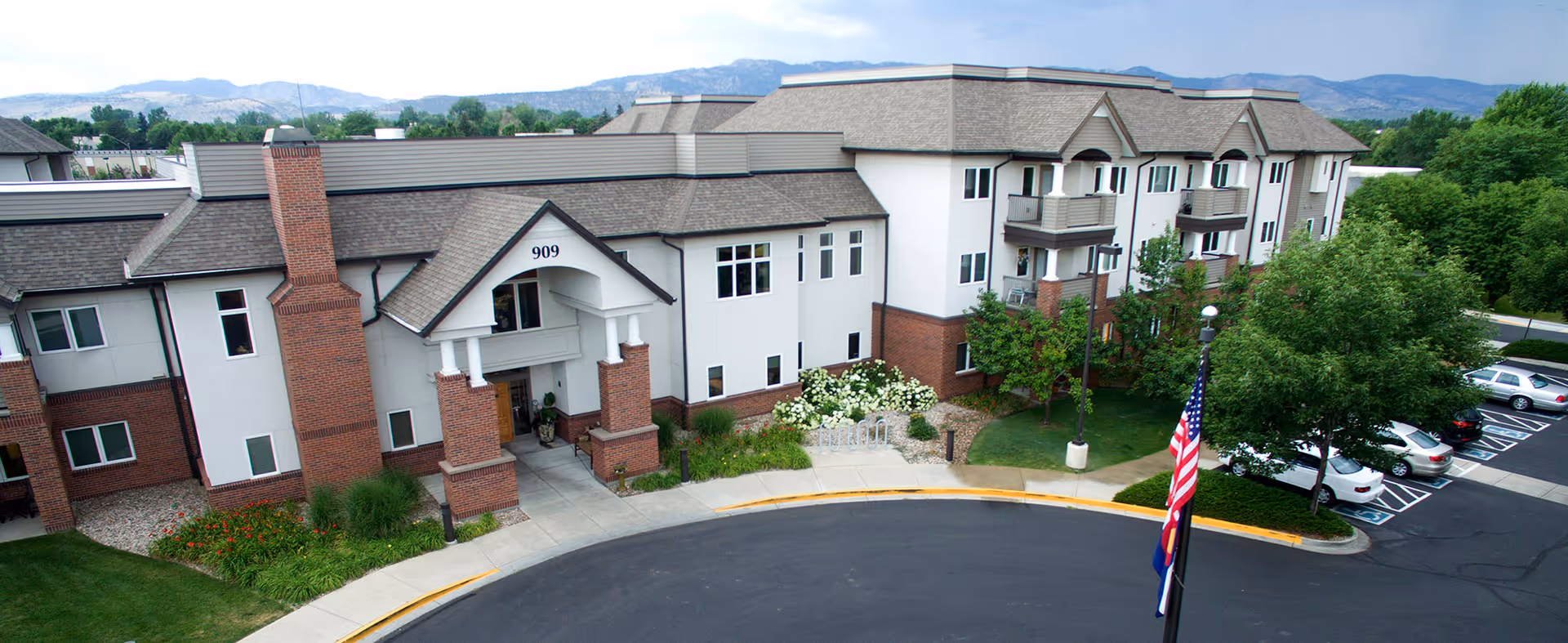 Exterior view of a multi-story senior living facility building with a brick and white facade, balconies, and a covered entrance marked with the number 909. There is a curved driveway in front, landscaped greenery, parked cars, and an American flag on a flagpole. Mountains and trees are visible in the background under a partly cloudy sky.