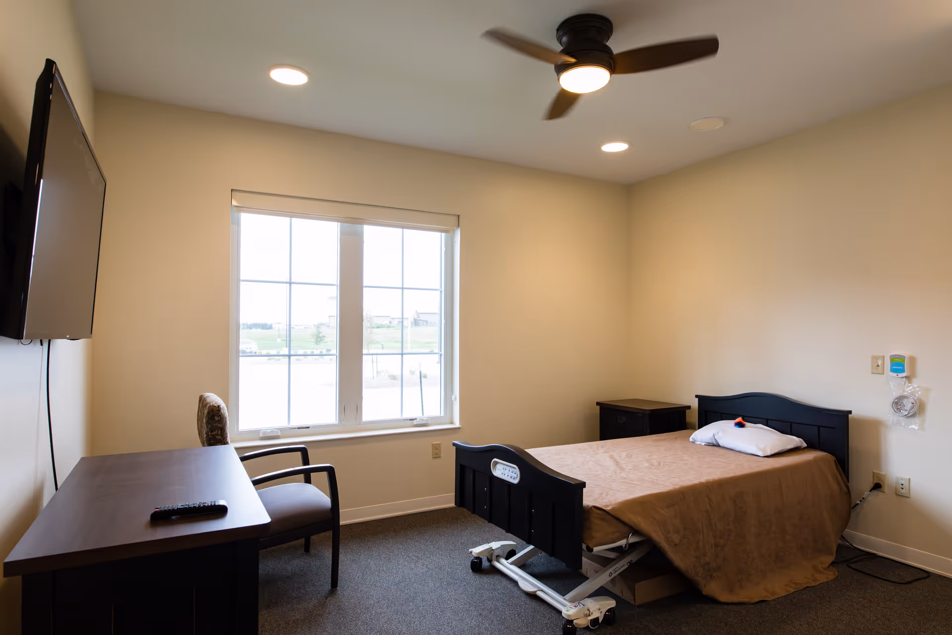 A simple senior living bedroom with a single adjustable bed covered with a brown blanket and a white pillow. The room has a window with a view outside, a ceiling fan with light, a wall-mounted TV, a wooden desk with a chair, and a small bedside table. The walls are painted beige and the floor is carpeted.