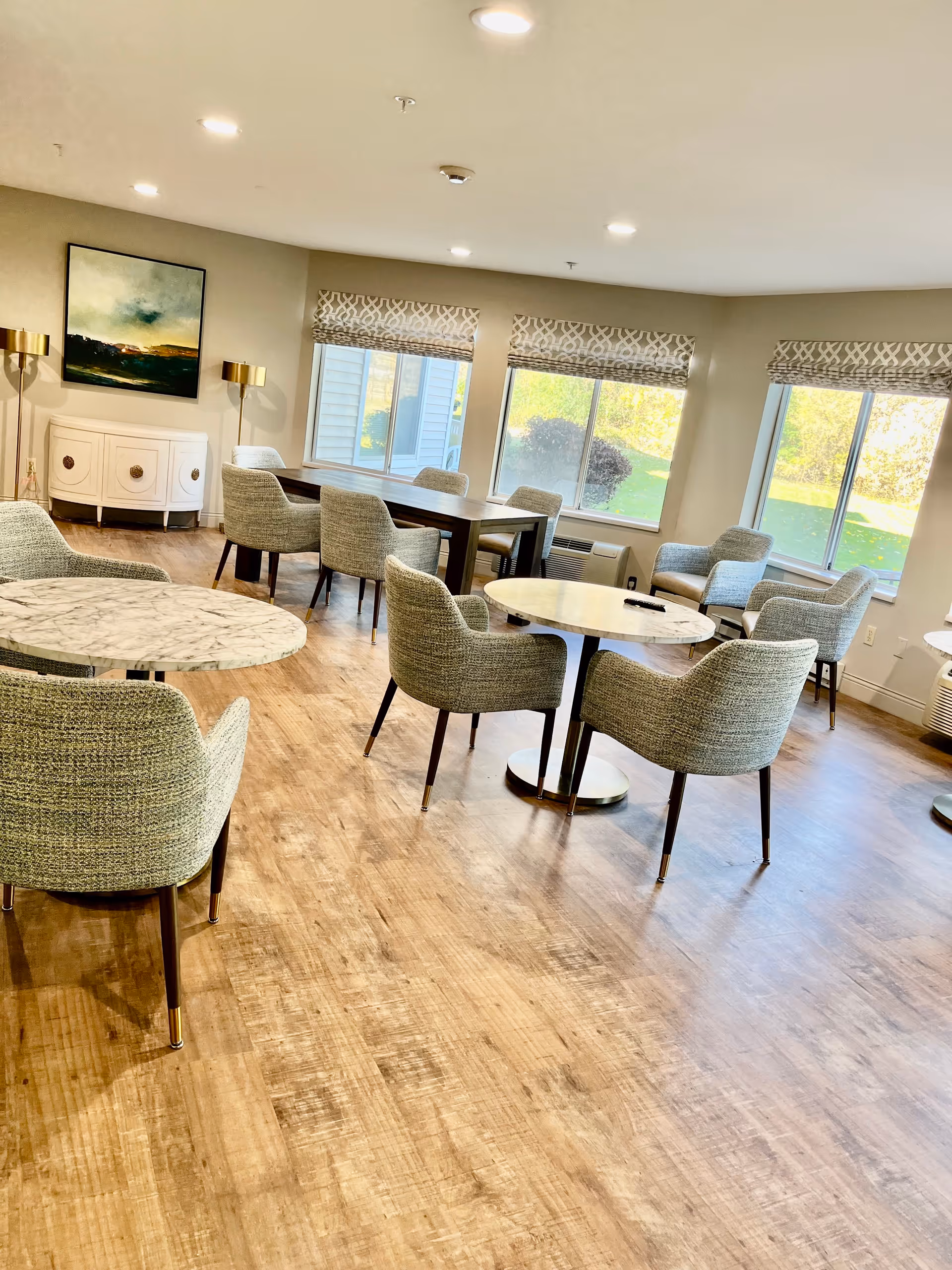 A bright and spacious common area with multiple round and rectangular tables surrounded by upholstered chairs. Large windows with patterned valances allow natural light to fill the room. The floor is wood, and there is a white sideboard with a painting and two gold floor lamps against one wall.