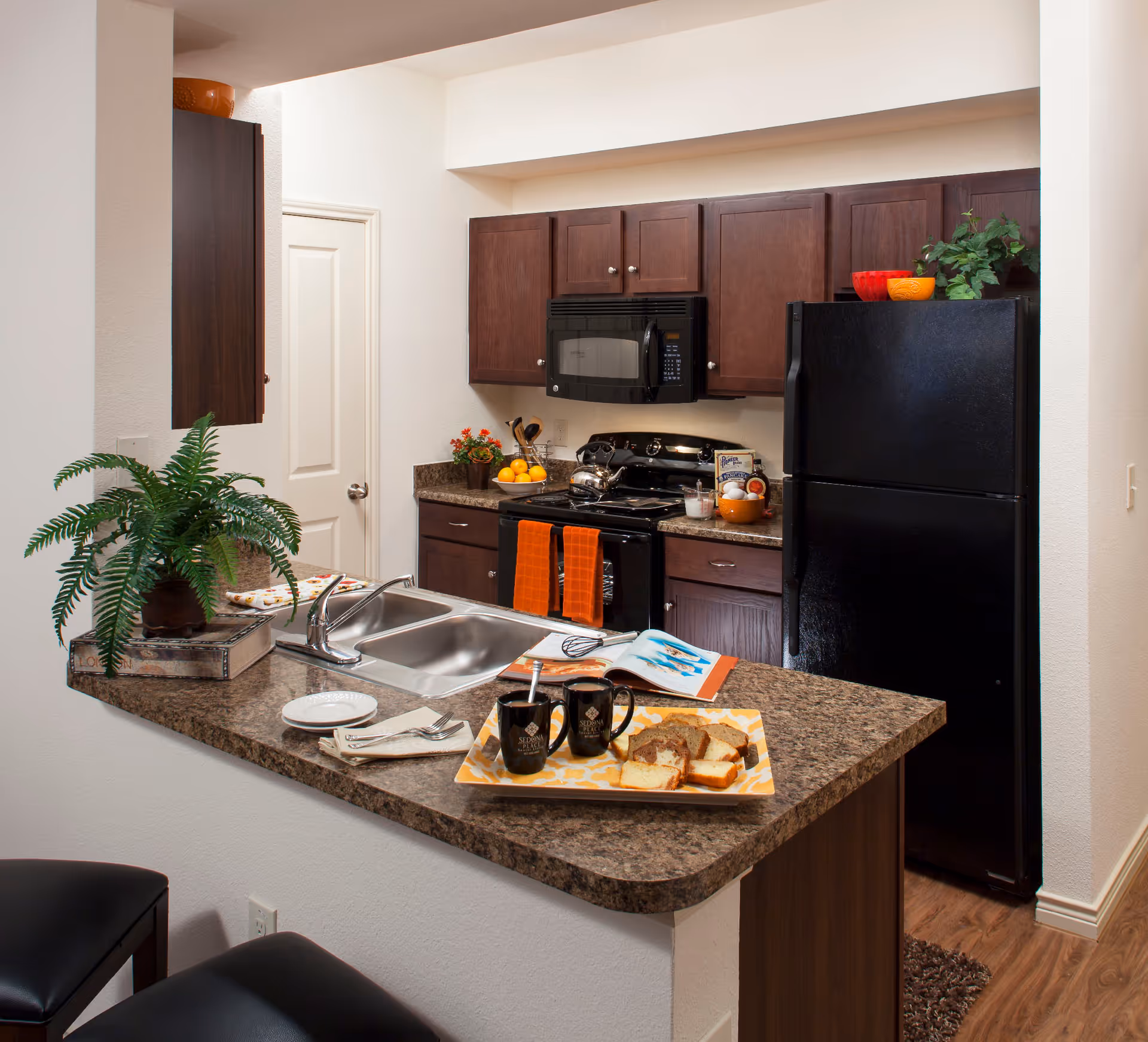 A modern kitchen with dark wood cabinets, a black refrigerator, black microwave, and black stove. The countertop has a double sink, a potted plant, two black mugs, a plate with sliced bread, and an open magazine. Two orange towels hang on the oven handle. The floor is wood, and there are two black stools at the counter.