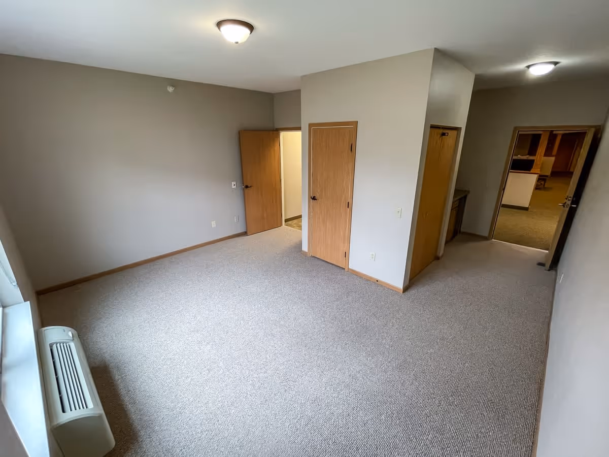 Empty room with beige carpet and light gray walls, featuring three wooden doors, a window with a heating/cooling unit below it, and ceiling light fixtures.