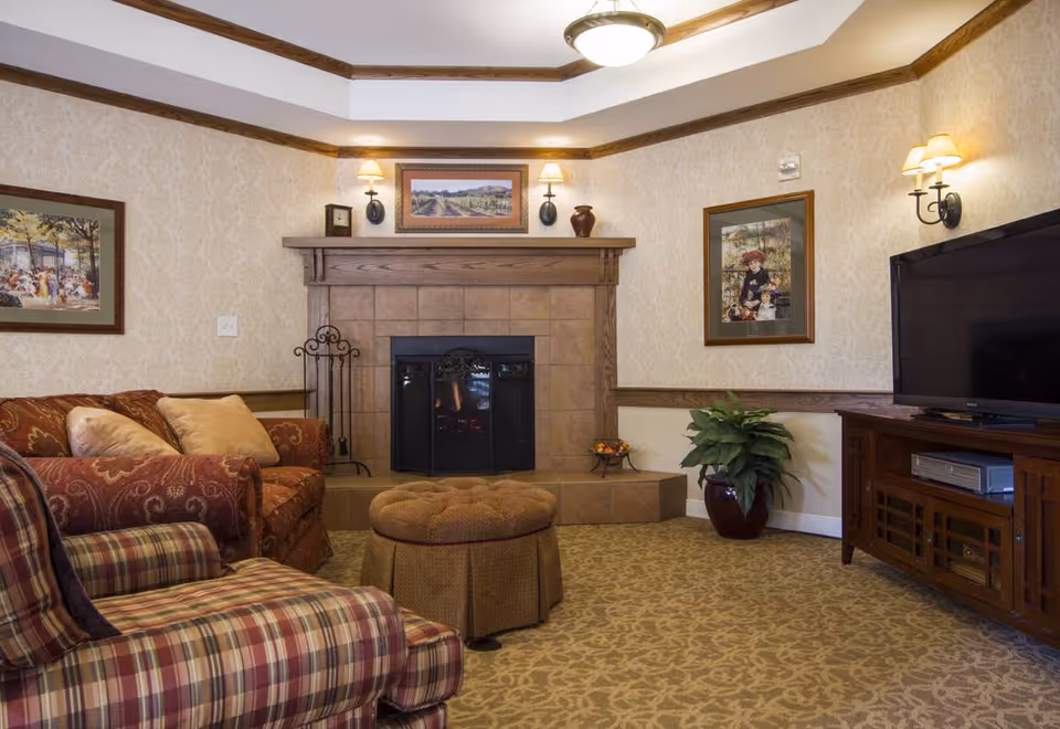 Cozy living room with a fireplace, upholstered chairs and an ottoman, a TV on a wooden console, and framed artwork on the walls.