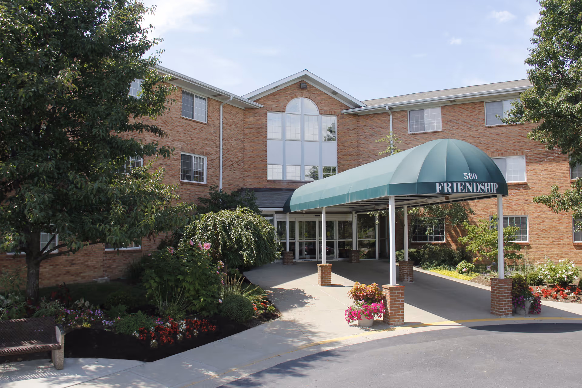 Exterior view of Friendship Towers, a three-story brick building with multiple windows. The entrance features a green canopy with the number 580 and the word 'FRIENDSHIP' on it. There are landscaped flower beds and trees around the entrance area.